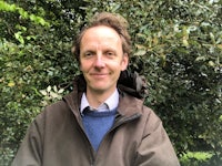 a man in a brown jacket standing in front of bushes